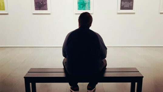 Mies istuu penkillä katsellen seinällä olevia taideteoksia.