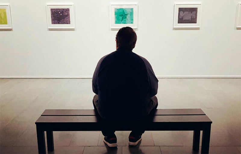 Mies istuu penkillä katsellen seinällä olevia taideteoksia.
