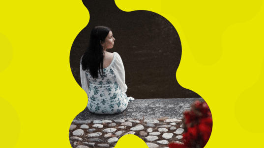 Nina istuu joenpenkalla selin kameraan.